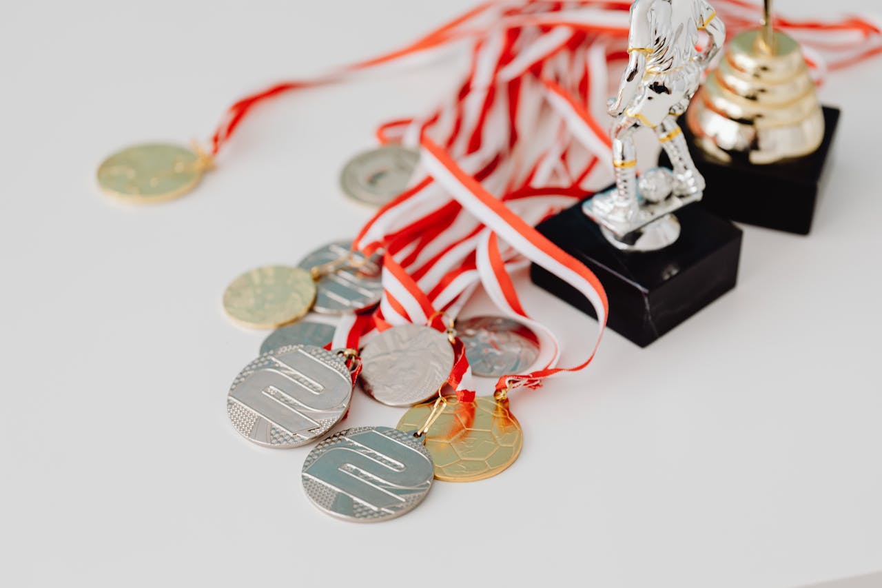 heros-img A varied collection of silver and gold medals alongside trophies on a white surface, showcasing achievement.